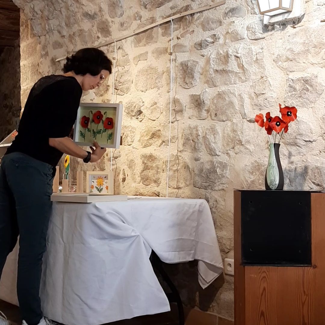 Installation d'une exposition - Galerie des Augustins - Puget-Theniers - Juillet 2024 Créatrice BéaCréations installant ses tableaux près d'une table avec des coquelicot en papier et un vase.